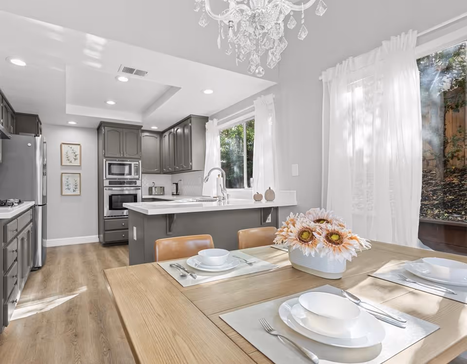 Bright and modern kitchen and dining area with gray cabinets, stainless steel appliances, a white countertop island with a sink, and a wooden dining table set with white dishes and a centerpiece of sunflowers. Large windows with sheer white curtains allow natural light to fill the space.