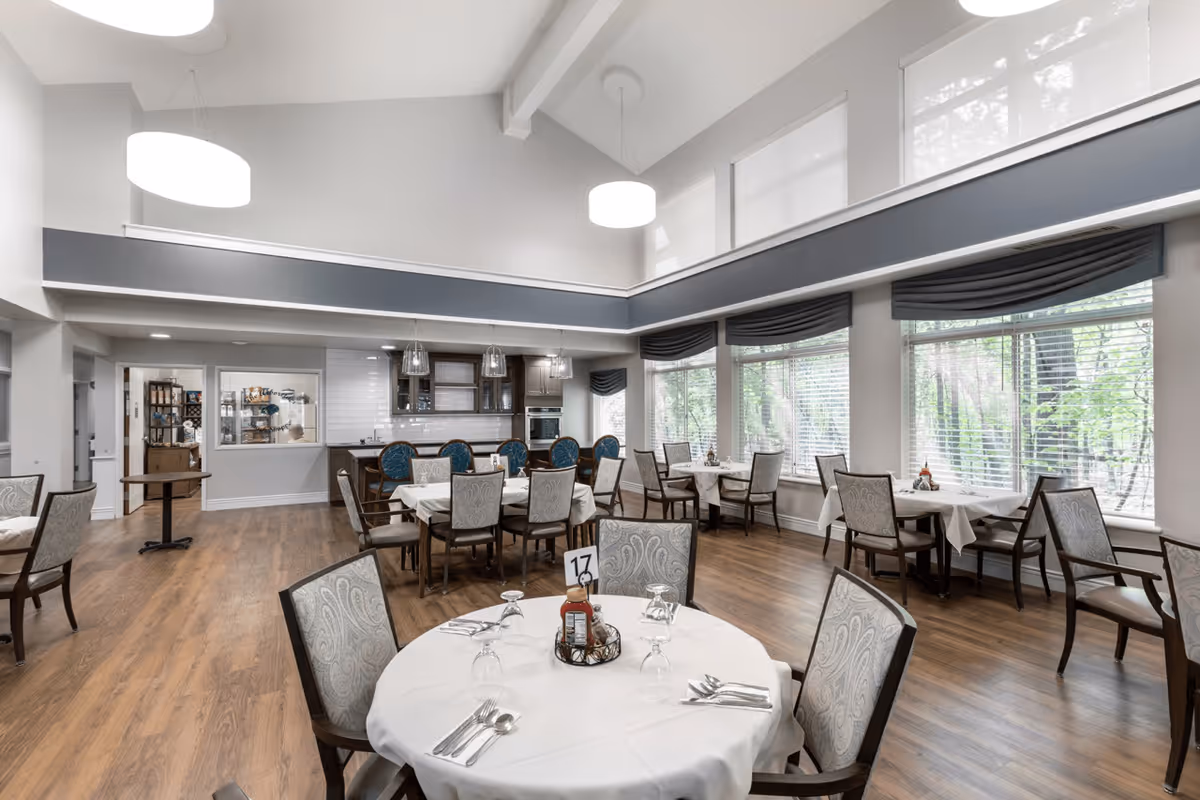 A bright and spacious dining room in a senior living facility with multiple round tables covered with white tablecloths, each set with silverware, glasses, and condiments. The room features large windows with blinds and valances, wooden flooring, and modern pendant lights hanging from a high ceiling. There is a kitchen area visible in the background with cabinetry and appliances.