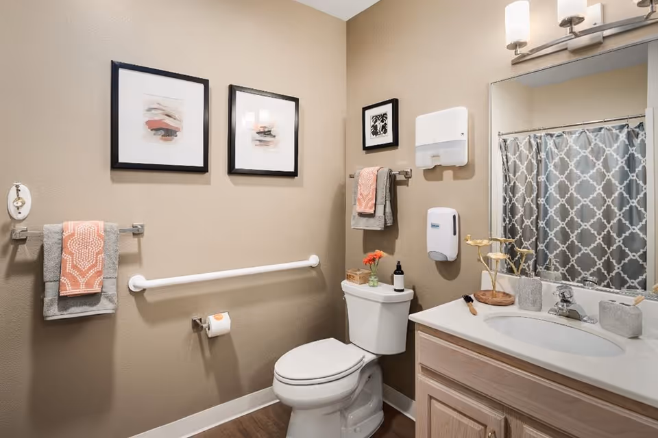 A clean and well-lit bathroom with beige walls, a white toilet, a white sink with a wooden cabinet, a large mirror above the sink, and a shower with a gray and white patterned curtain. There are two framed abstract artworks on the wall above the toilet, a towel rack with gray and coral towels, a hand sanitizer dispenser, a paper towel dispenser, and a grab bar next to the toilet.