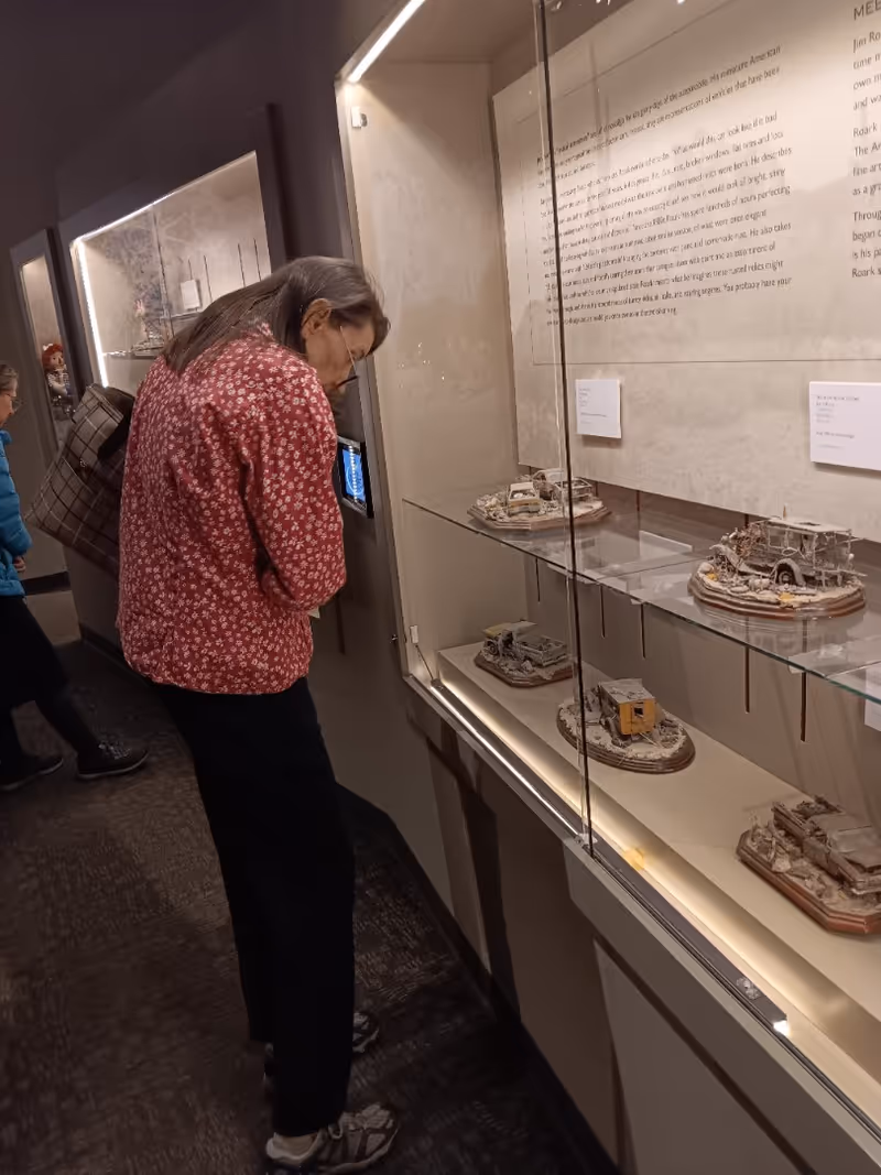 An elderly woman wearing a red floral jacket and black pants is closely observing miniature models displayed in a glass case at a museum or exhibit. Another person is visible in the background also looking at displays along the wall.