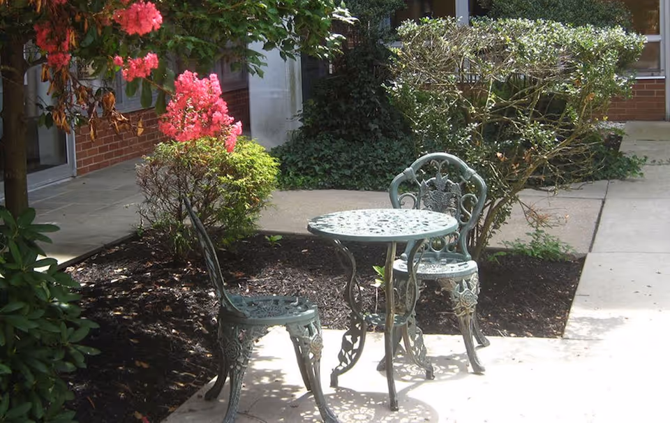 A small outdoor patio area with two ornate metal chairs and a matching round table. The patio is surrounded by plants and bushes, including a flowering bush with bright pink flowers. A brick building and windows are visible in the background.