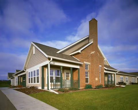 Exterior view of a single-story senior living facility building with beige siding, green trim, multiple windows, a prominent brick chimney, and a covered porch area. The building is surrounded by a well-maintained lawn and a concrete walkway under a partly cloudy blue sky.