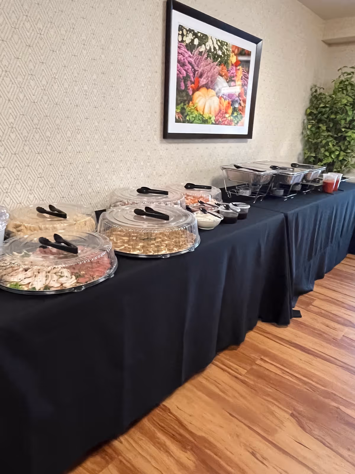A long buffet table with covered platters and chafing dishes set against a patterned wall with framed artwork.