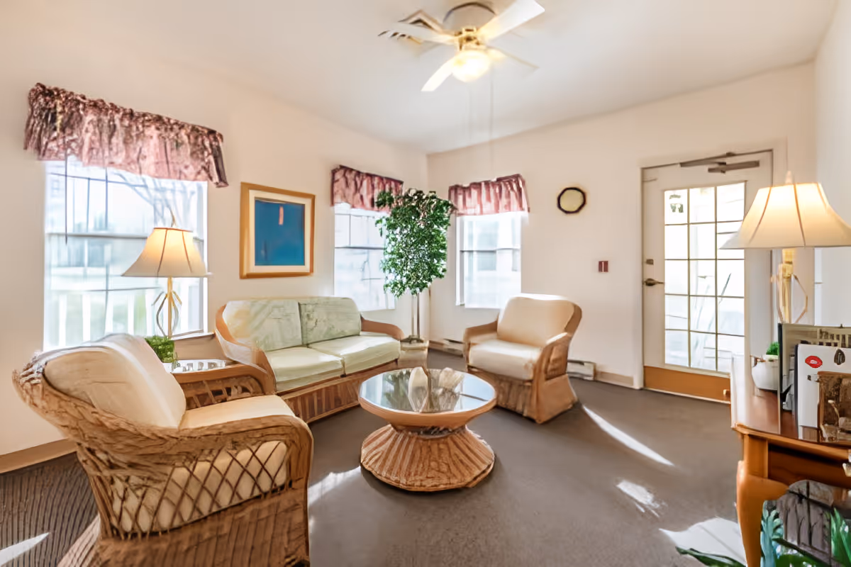 A bright living room with wicker furniture including a sofa and two armchairs with light cushions, a round glass-top coffee table, two table lamps, a potted plant, three windows with pink valances, a ceiling fan, and a door with glass panels letting in natural light.