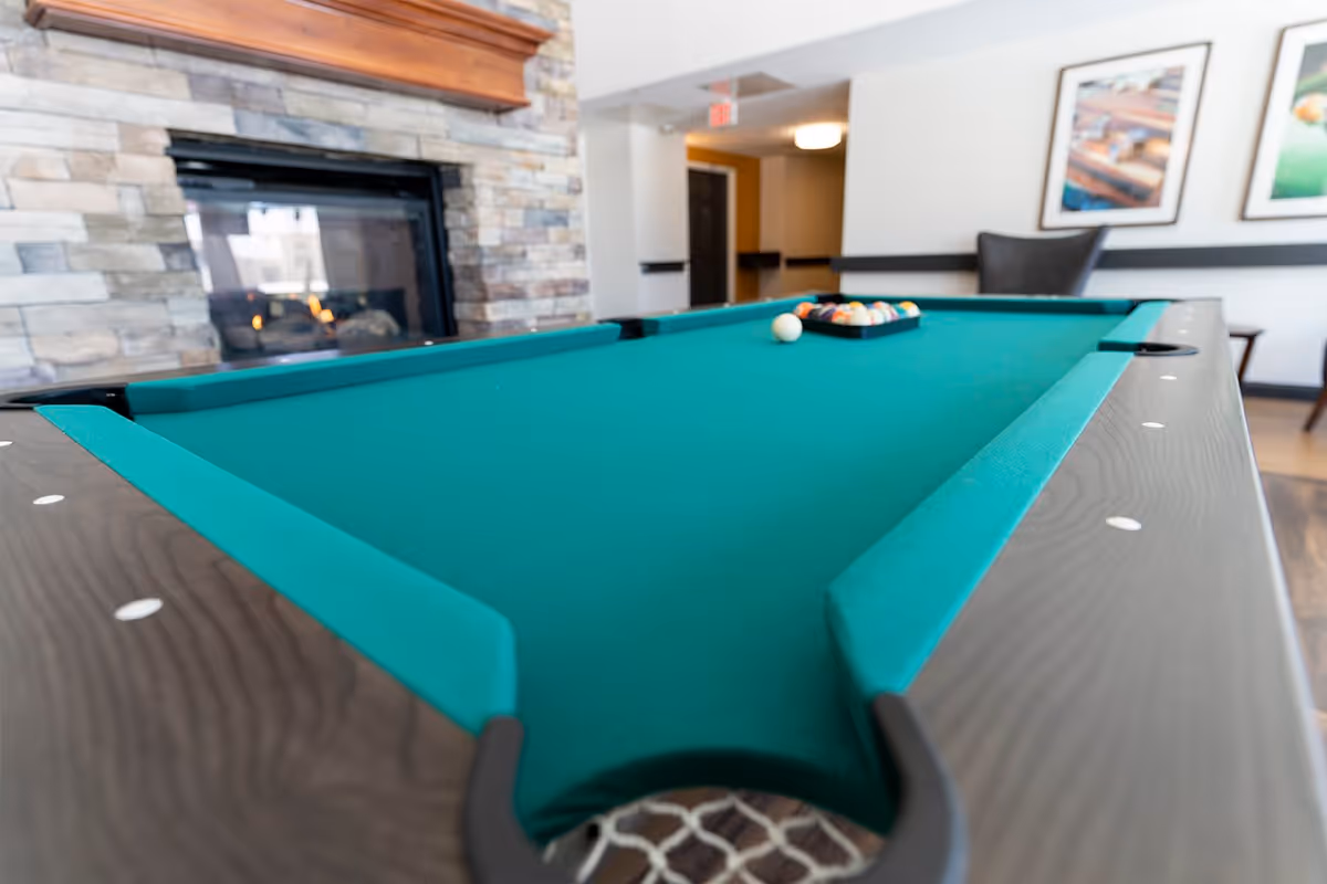 A green-felt pool table in a lounge area with a stone fireplace and seating in the background.