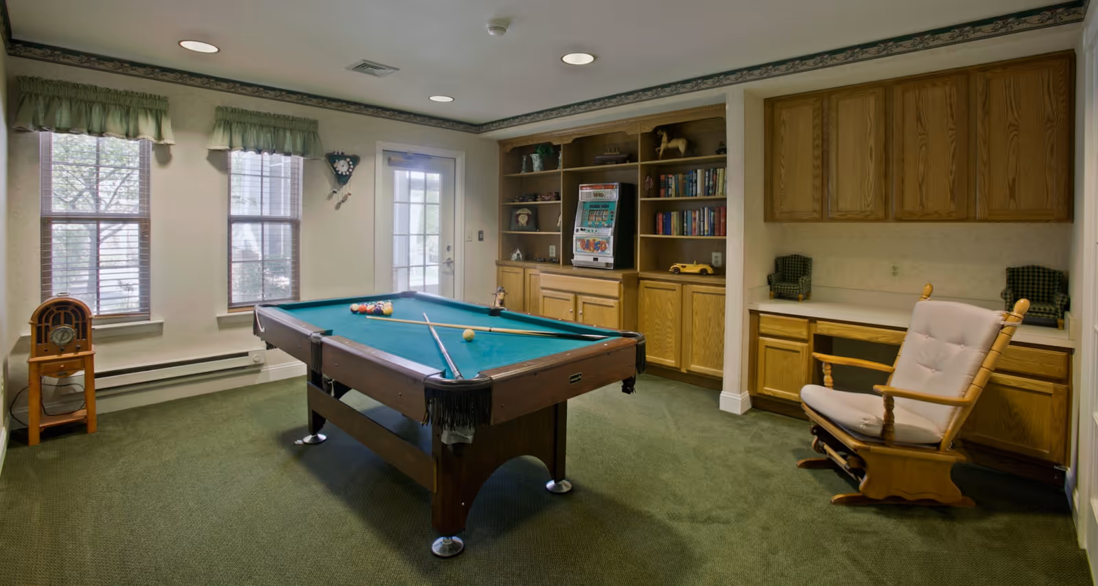 A recreational room with a pool table in the center, two pool cues resting on it, and a set of billiard balls arranged. The room has green carpet, two windows with green valances, a door with glass panes, built-in wooden shelves and cabinets filled with books and decorative items, and a cushioned wooden rocking chair near a desk area.