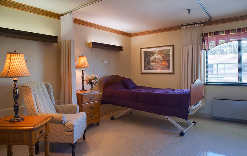 Furnished nursing facility bedroom with an adjustable single bed covered in a purple blanket, an armchair, bedside tables with lamps and a window.