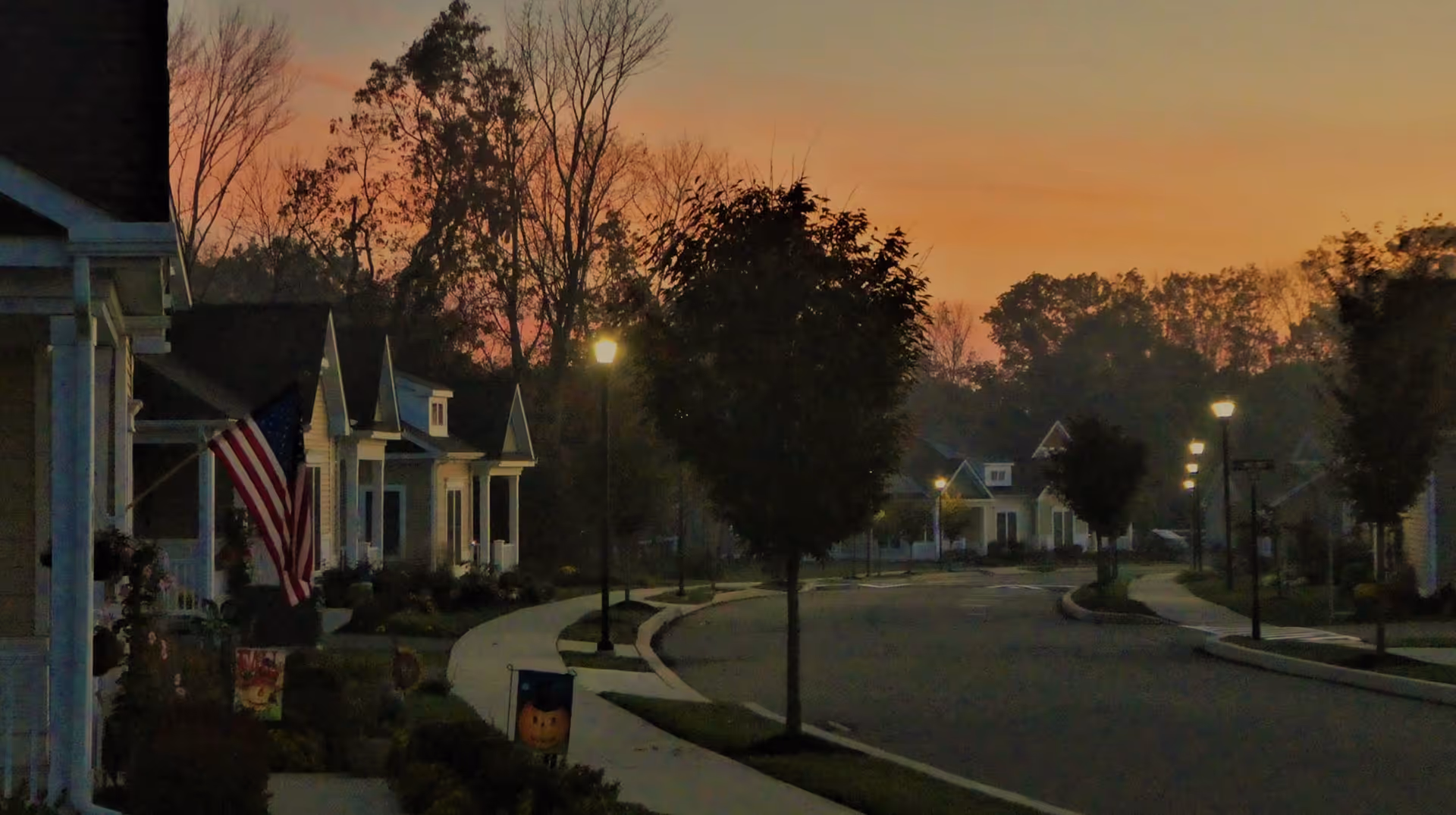A quiet residential street at dusk with small houses lined along the curved road. Trees and street lamps are visible, with a soft orange and purple sky in the background. An American flag is displayed on one of the houses.