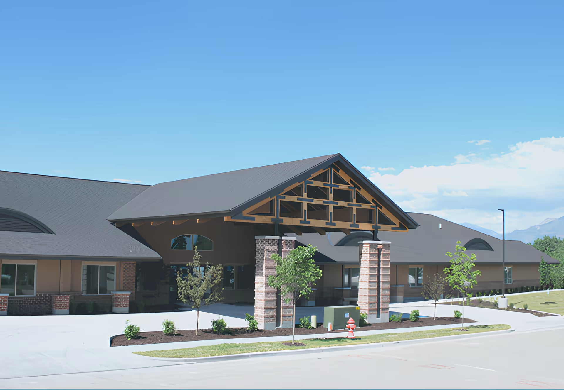 Front entrance of a senior living building with a covered porte-cochere, stone columns, small trees and landscaping under a clear blue sky.