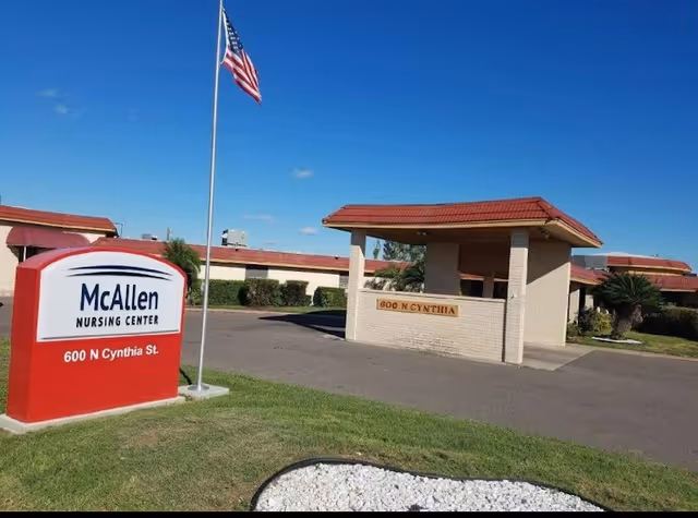 Exterior view of McAllen Nursing Center showing the main entrance with a covered drop-off area, a red and white sign displaying the facility name and address, an American flag on a flagpole, and a clear blue sky.