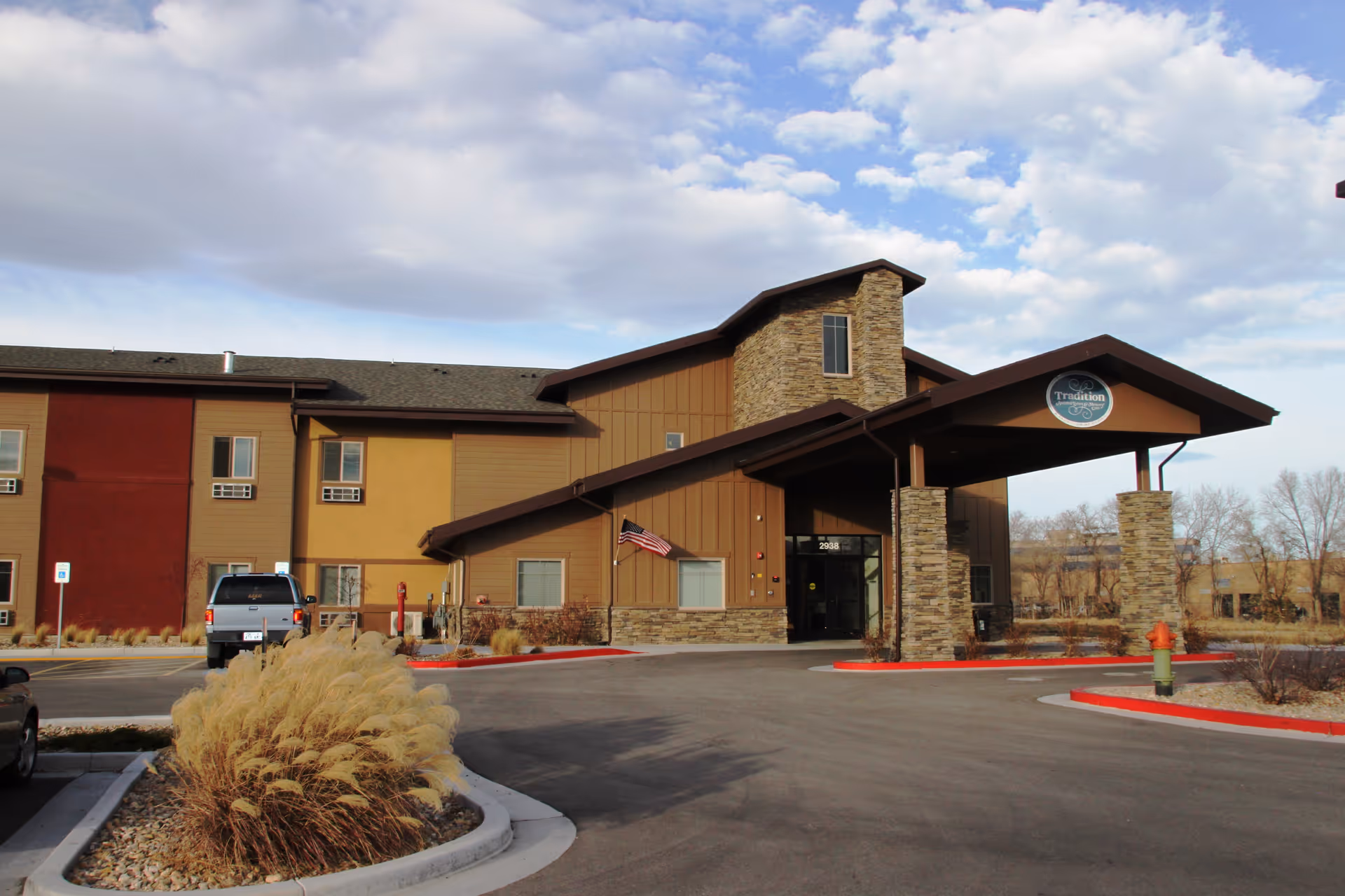 Exterior view of Tradition Assisted Living facility showing a two-story building with a covered entrance supported by stone pillars, an American flag near the entrance, and a parking area with some vehicles. The sky is partly cloudy.