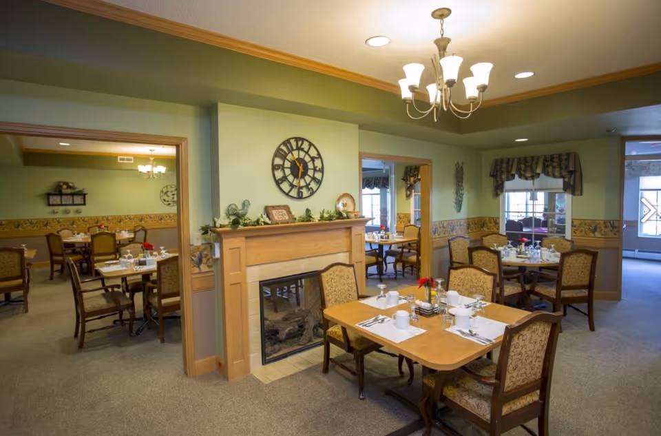 A cozy dining room in a senior living facility with multiple wooden tables and upholstered chairs set with white cups, plates, and silverware. The room features a decorative fireplace with a clock above it, green walls with a floral border, and a chandelier hanging from the ceiling. There are windows with valances and an adjoining dining area visible through open doorways.