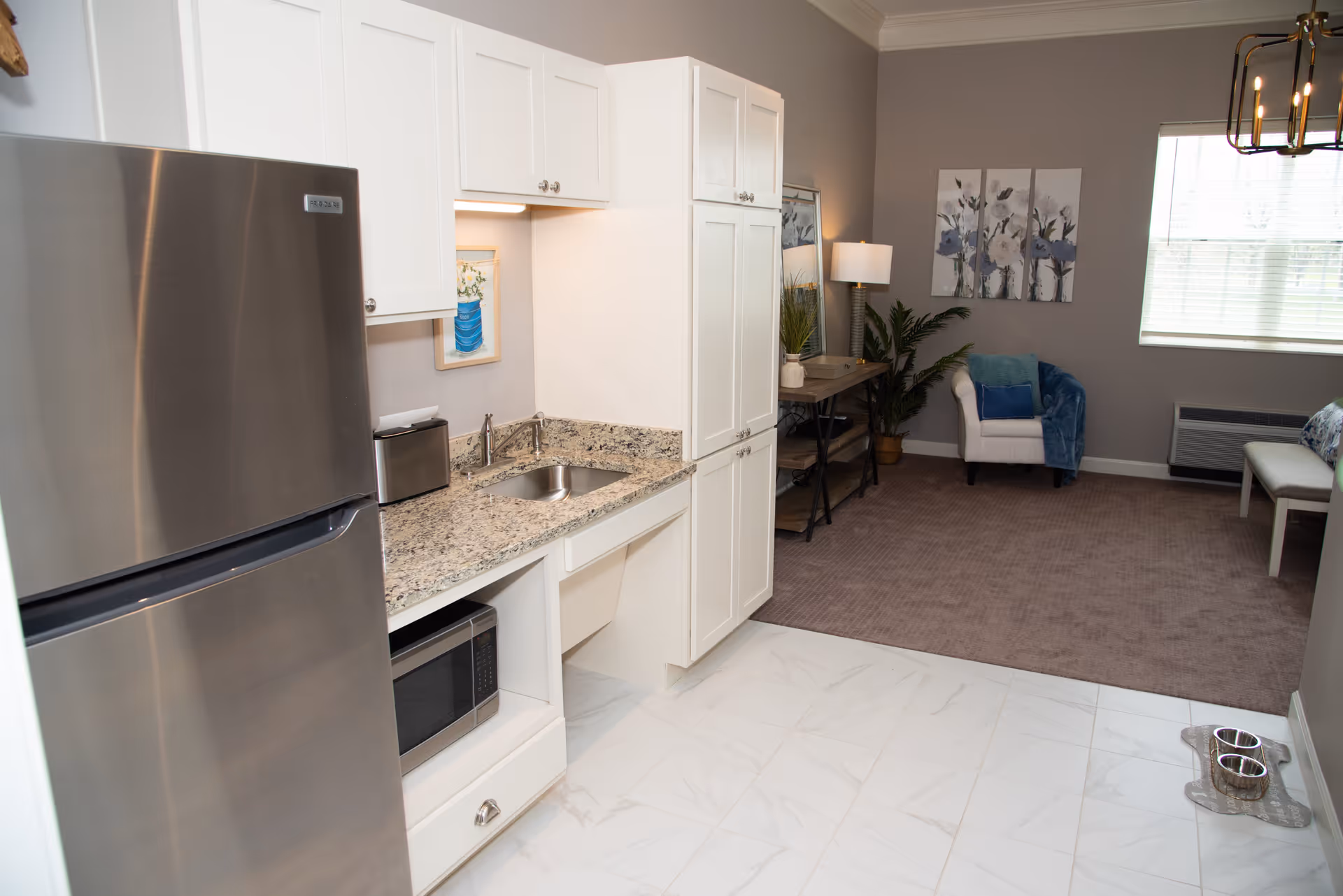Open studio kitchenette with a stainless refrigerator, granite countertop and sink opening into a carpeted living area with an armchair and window.