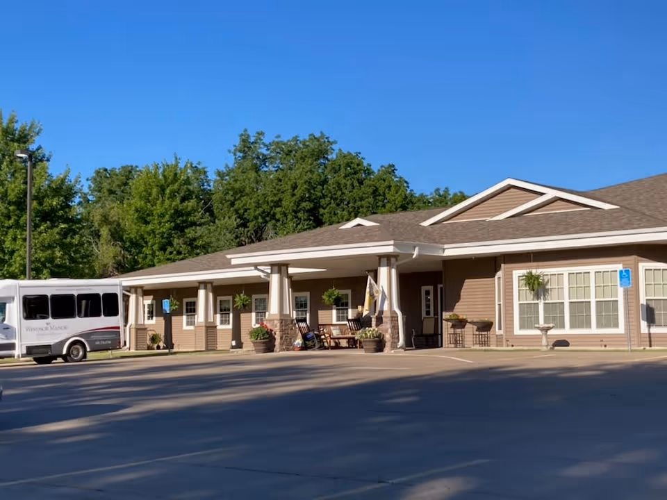 Exterior front of Windsor Manor: a single-story senior living building with a covered entrance, parked shuttle van, planters, and trees under a clear blue sky.