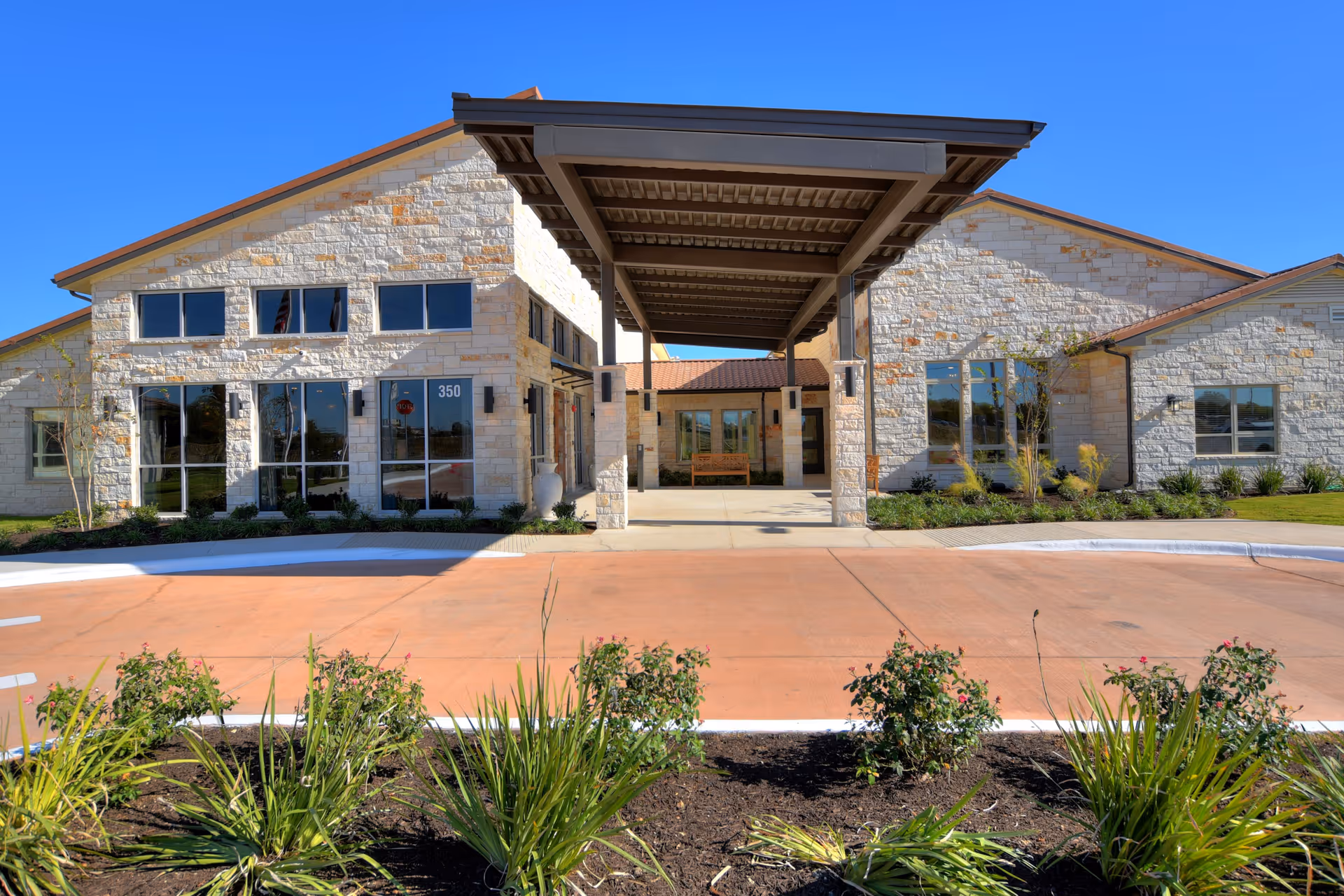 Front exterior view of The Philomena facility showing a stone building with large windows, a covered entrance with pillars, a driveway, and landscaping with plants and bushes under a clear blue sky.