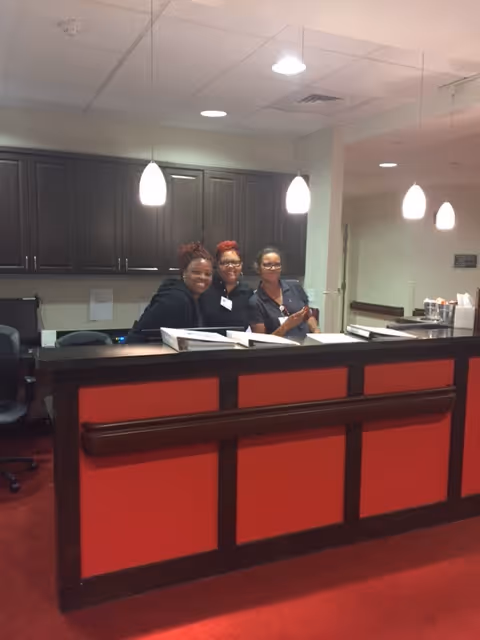 Three staff members smiling behind a red reception desk in a senior living facility lobby.
