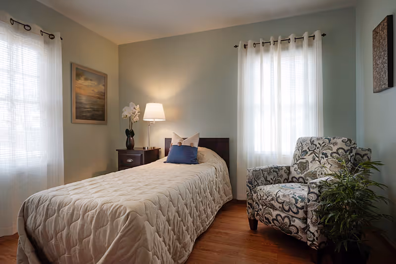 A cozy bedroom with a single bed covered in a beige quilted bedspread and two pillows. Next to the bed is a wooden nightstand with a lamp and a vase with white flowers. There is a patterned armchair beside a green potted plant near a window with sheer white curtains. The walls are painted light green and decorated with framed artwork.