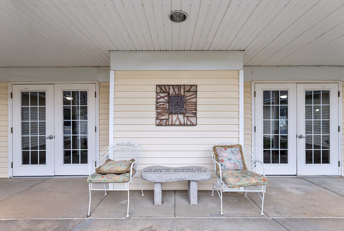 Outdoor covered patio area with two white metal chairs with floral cushions and a small stone memorial bench between them. The wall behind has beige siding and a decorative clock with Roman numerals. There are two sets of white double doors with glass panes on either side of the seating area.