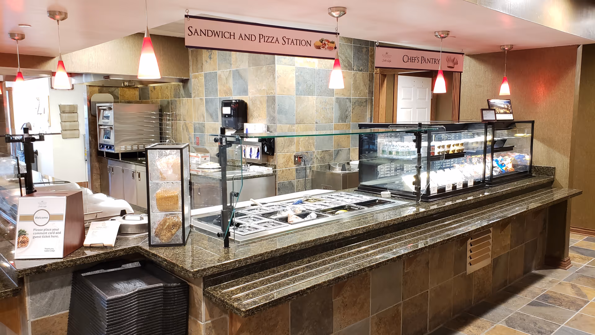Interior view of a food service counter at Sable Lodge Retirement Community featuring a sandwich and pizza station with various food items behind a glass barrier. The area has tiled walls and floors, pendant lights hanging from the ceiling, and signs indicating 'Sandwich and Pizza Station' and 'Chef's Pantry'.