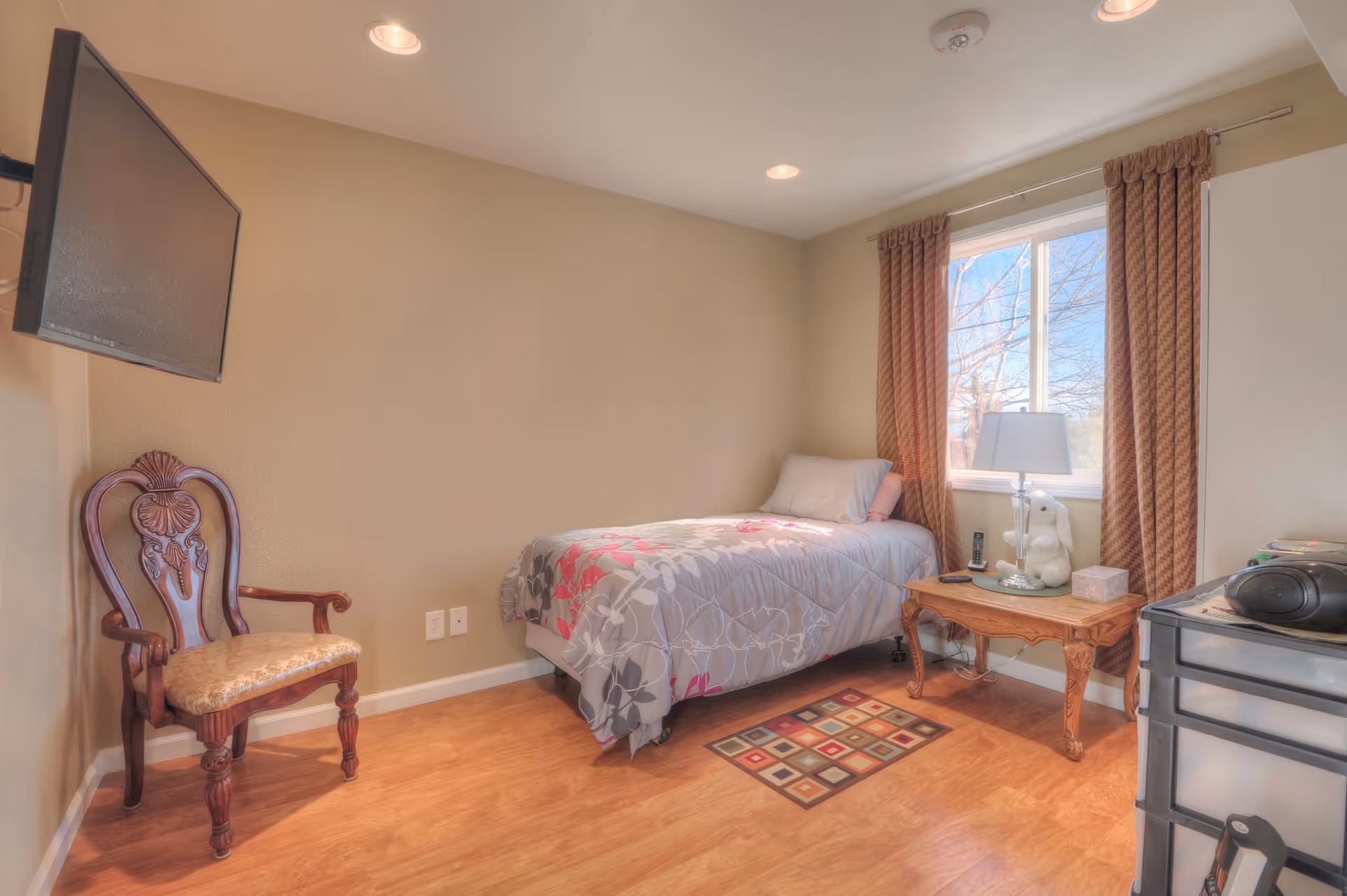 A small bedroom with a single bed covered in a floral-patterned comforter, a wooden chair with an upholstered seat, a wall-mounted flat-screen TV, a wooden side table with a lamp and a stuffed animal, a window with brown curtains, and a plastic drawer unit on the right side.