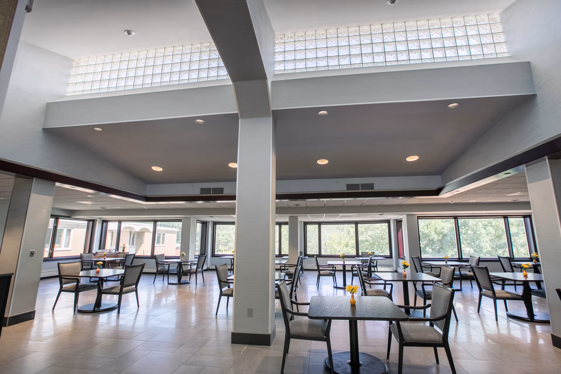 A spacious dining room with multiple square tables and chairs arranged neatly. Each table has a small vase with yellow flowers. Large windows line the walls, allowing natural light to fill the room. The ceiling features recessed lighting and a high section with glass blocks for additional light.