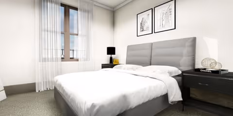 A modern bedroom with a large bed featuring a gray upholstered headboard and white bedding. There are two black nightstands on either side of the bed, one with a black lamp and a yellow book, and the other with a decorative globe and a Chanel book. The room has a window with sheer white curtains and two framed black and white artworks hanging above the bed.