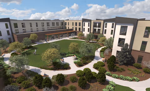 A large, modern three-story senior living facility building surrounding a spacious landscaped courtyard with green lawns, walking paths, trees, shrubs, and benches. Several people are seen walking and sitting in the courtyard area under a partly cloudy sky.