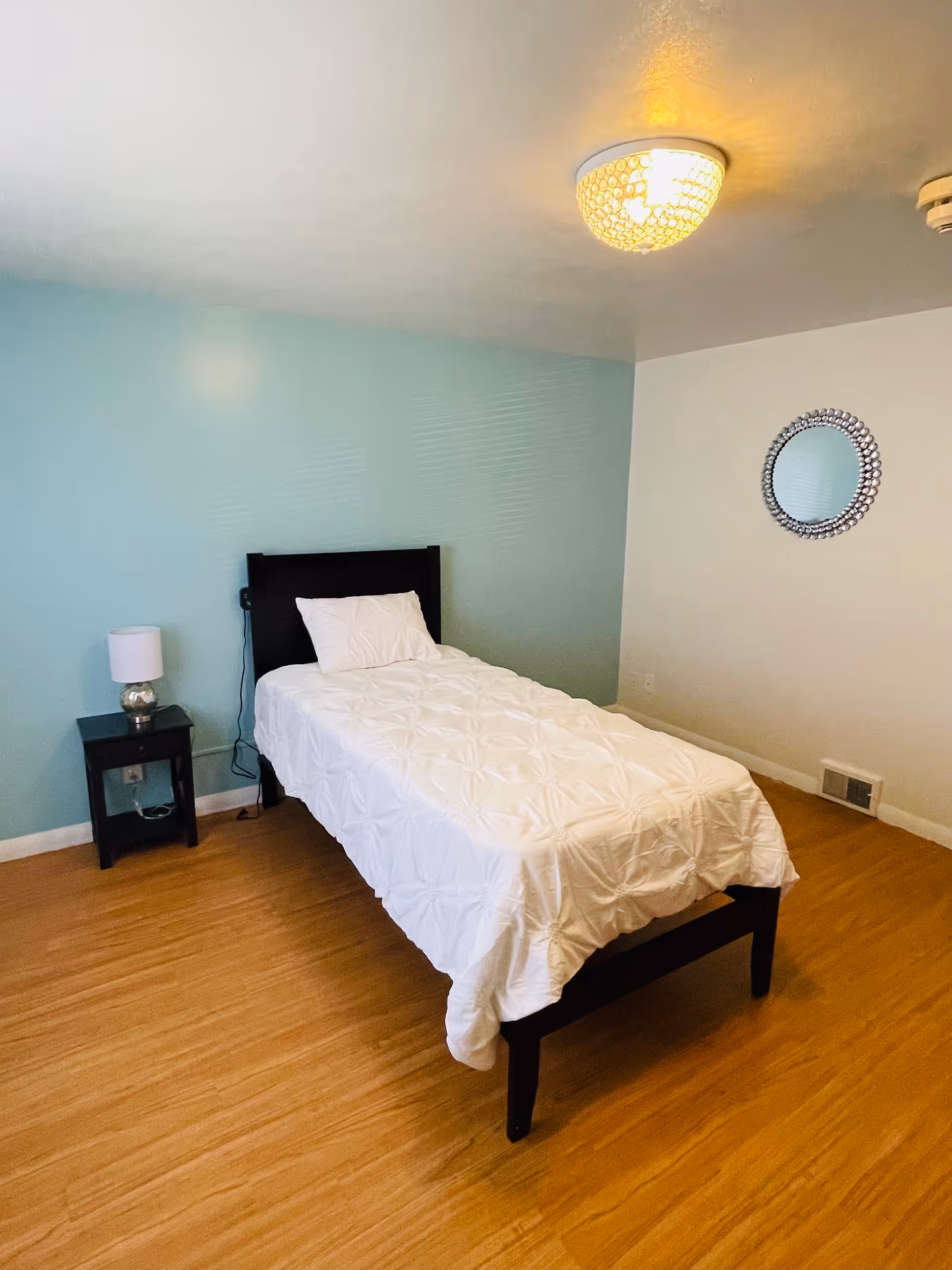 A simple bedroom with a single bed covered in a white textured comforter and a white pillow. The bed has a dark wooden headboard. Next to the bed is a small black nightstand with a silver lamp. The walls are painted light blue and white, and there is a round decorative mirror on the white wall. The floor is wooden, and a ceiling light fixture is visible.