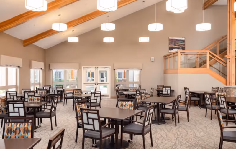Spacious senior living dining room with multiple tables and chairs, high ceilings with pendant lights, and large windows.