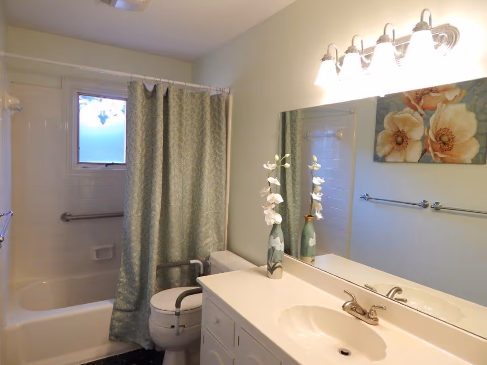 A bathroom with a white bathtub and shower combination, a green patterned shower curtain, a toilet with safety rails, a white vanity with a sink, a large mirror above the sink, a vase with white flowers on the counter, a towel rack on the wall, and a painting of large flowers. The bathroom has light-colored walls and a window above the bathtub.