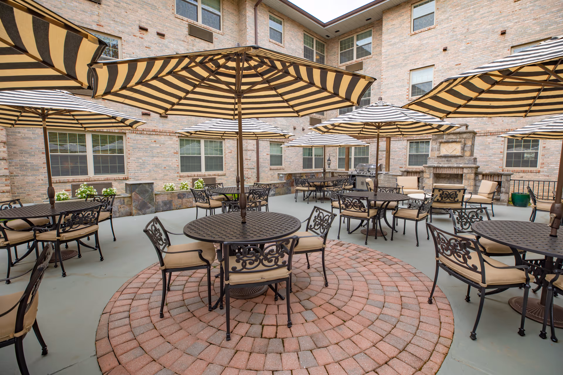 Outdoor patio area at Heatherwood Retirement Community with multiple round tables and chairs, each shaded by large striped umbrellas. The patio is surrounded by a brick building with several windows, and there is a stone fireplace and grill area in the background.