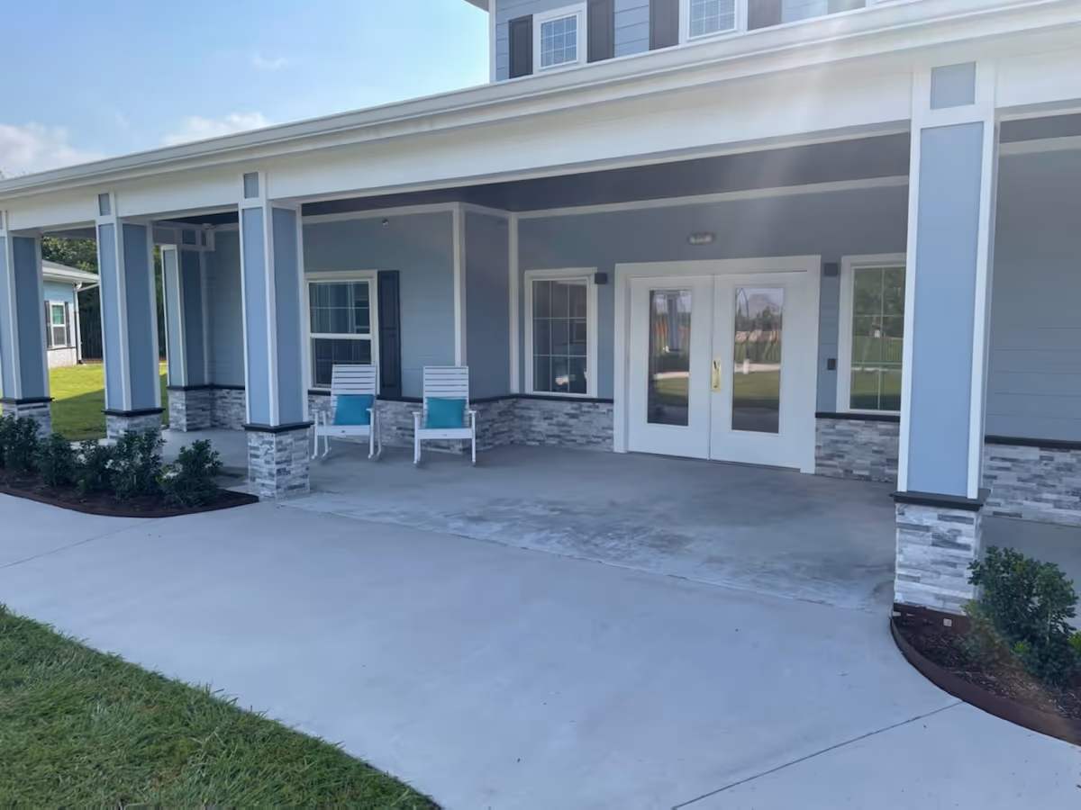 Covered front entrance of a light-blue building with stone-accent columns, two white chairs with teal cushions, and double glass doors.