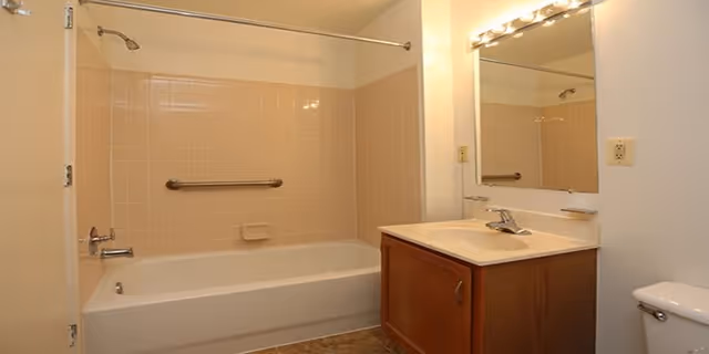 Bathroom with a bathtub and shower combination featuring beige tiles and a metal grab bar. There is a wooden vanity with a sink and a large mirror above it. A toilet is partially visible on the right side.