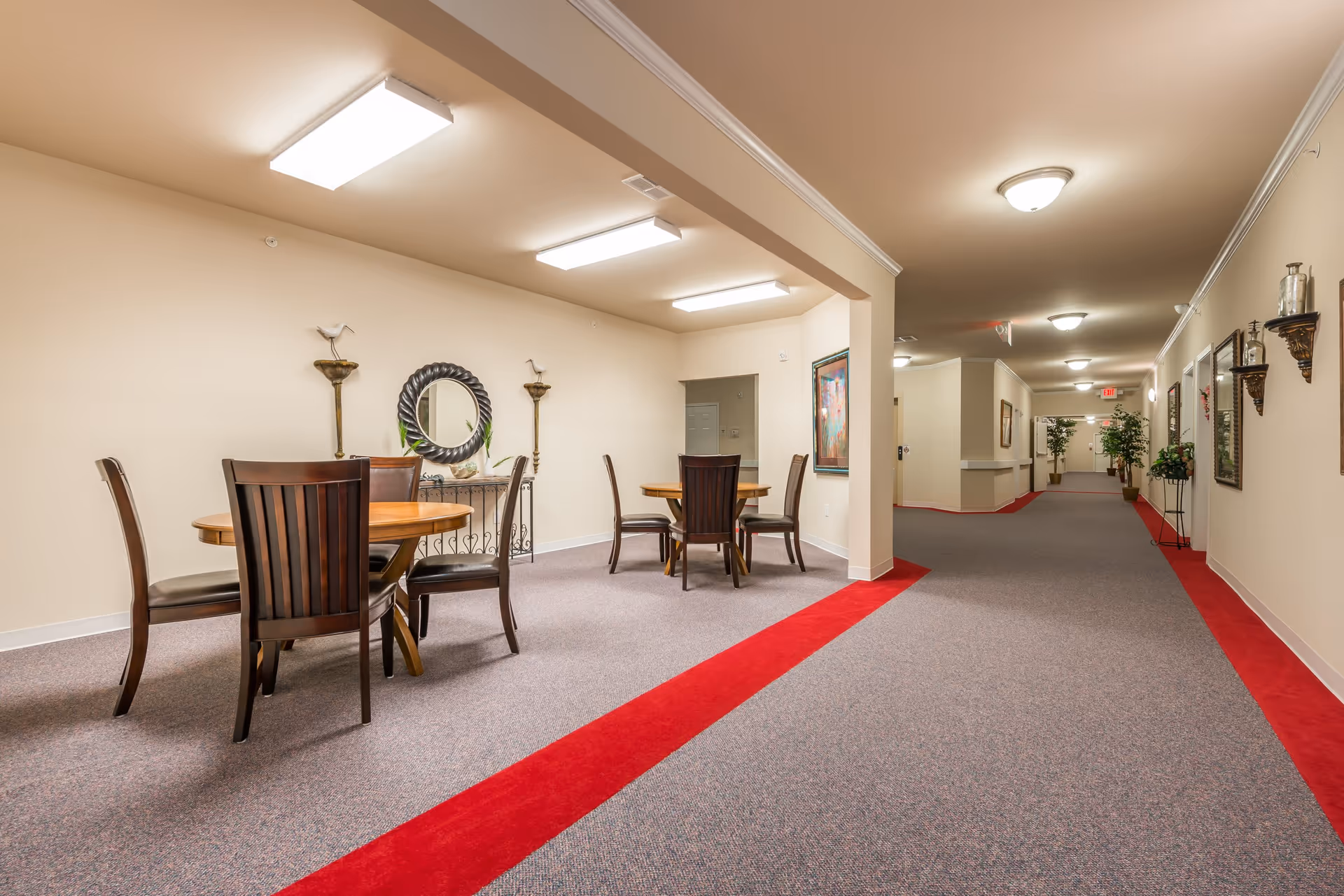 Interior view of a senior living facility hallway with a seating area on the left featuring two round wooden tables and several wooden chairs. The hallway has beige walls, a carpet with a red border, ceiling lights, wall decorations including a round mirror, sconces, and framed artwork, and potted plants along the corridor.