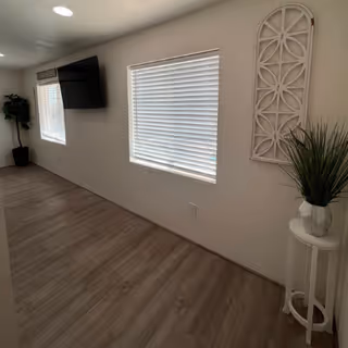 A hallway with light wood flooring and white walls featuring two windows with closed blinds. A flat-screen TV is mounted on the wall near one window. There is a tall potted plant in the corner and a small white round table with a decorative plant and wall art above it on the right side.