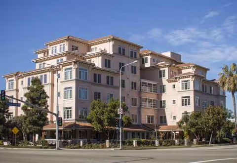 A multi-story senior living building is viewed from the street with trees and a clear sky.
