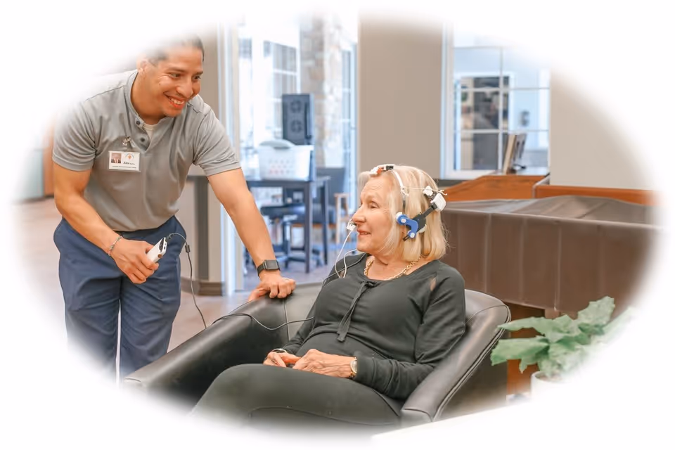 A smiling male caregiver or technician is assisting an elderly woman seated in a black chair. The woman is wearing a medical or therapeutic device on her head with wires attached, and they are in a bright, modern indoor setting with large windows and some furniture in the background.
