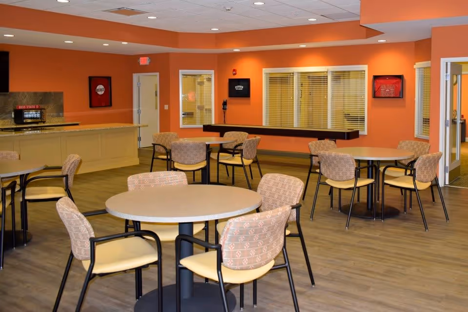 A dining area with several round tables and cushioned chairs arranged around them. The walls are painted orange, and there is a counter with a microwave on it. The floor is wood-style, and there are windows with blinds and framed pictures on the walls.