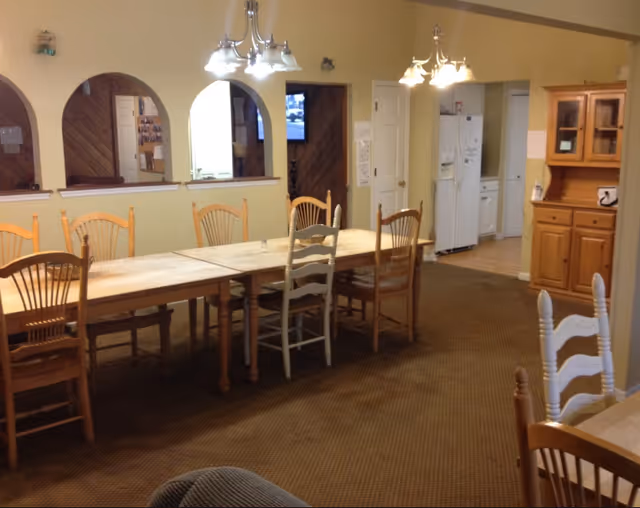 Long wooden dining table with mismatched chairs in a communal dining room, overhead lights, and a kitchenette/hutch area in the background.