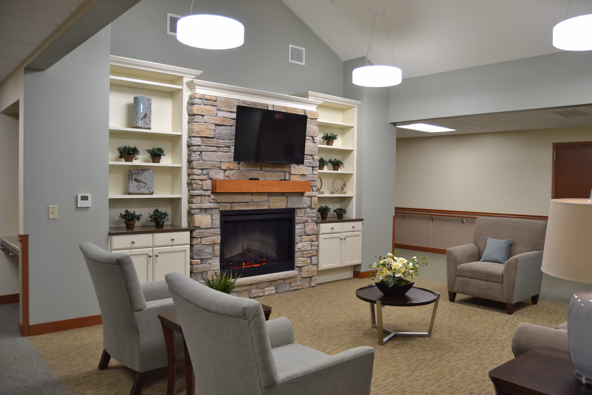 A cozy living room area in an assisted living facility featuring a stone fireplace with a mounted flat-screen TV above it. The room has light green walls, built-in white shelves with small potted plants and decorative items, and beige carpet. There are three upholstered armchairs arranged around a small round coffee table with a flower arrangement. Modern pendant lights hang from the ceiling.