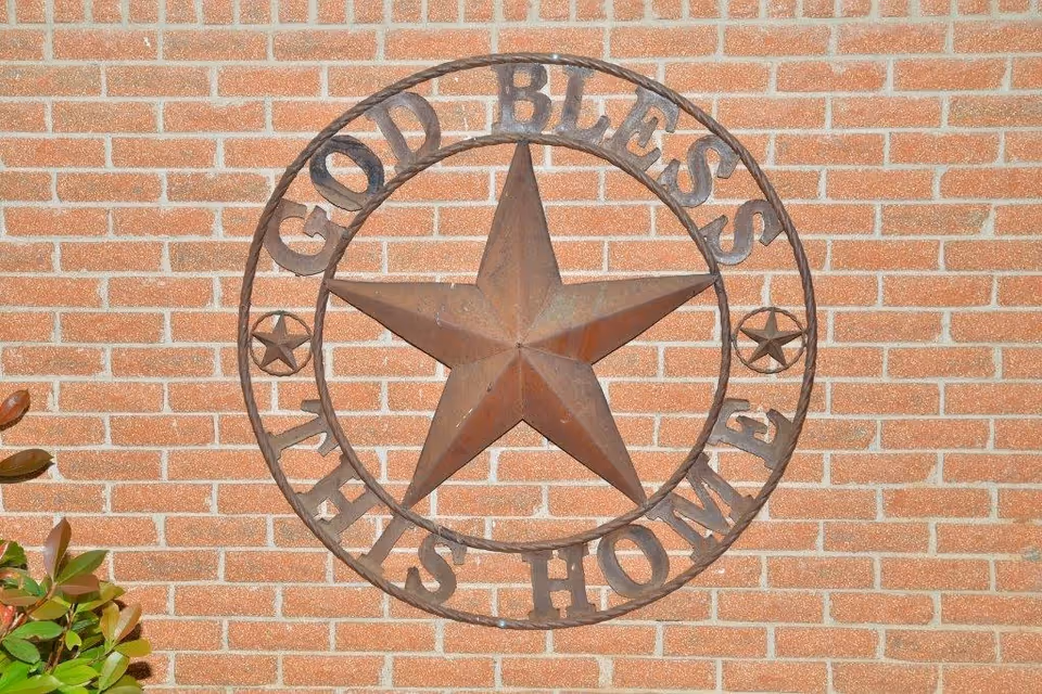 Rusty metal five-point star in a circular frame with the words "GOD BLESS TEXAS HOME" mounted on a brick exterior wall with a few leaves at the lower left.