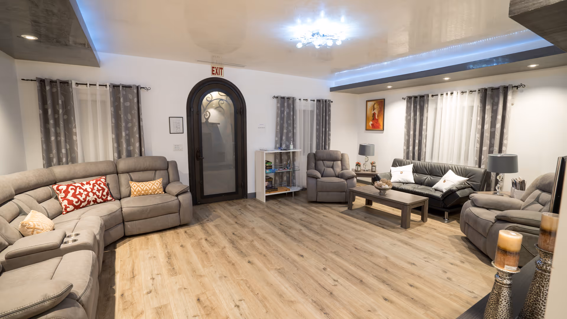 A spacious and well-lit senior living common area with light wood flooring, gray sectional and recliner sofas, a black leather couch, a wooden coffee table, side tables with lamps, decorative pillows, curtains on windows, a glass door with an exit sign above, and a small shelf with board games.