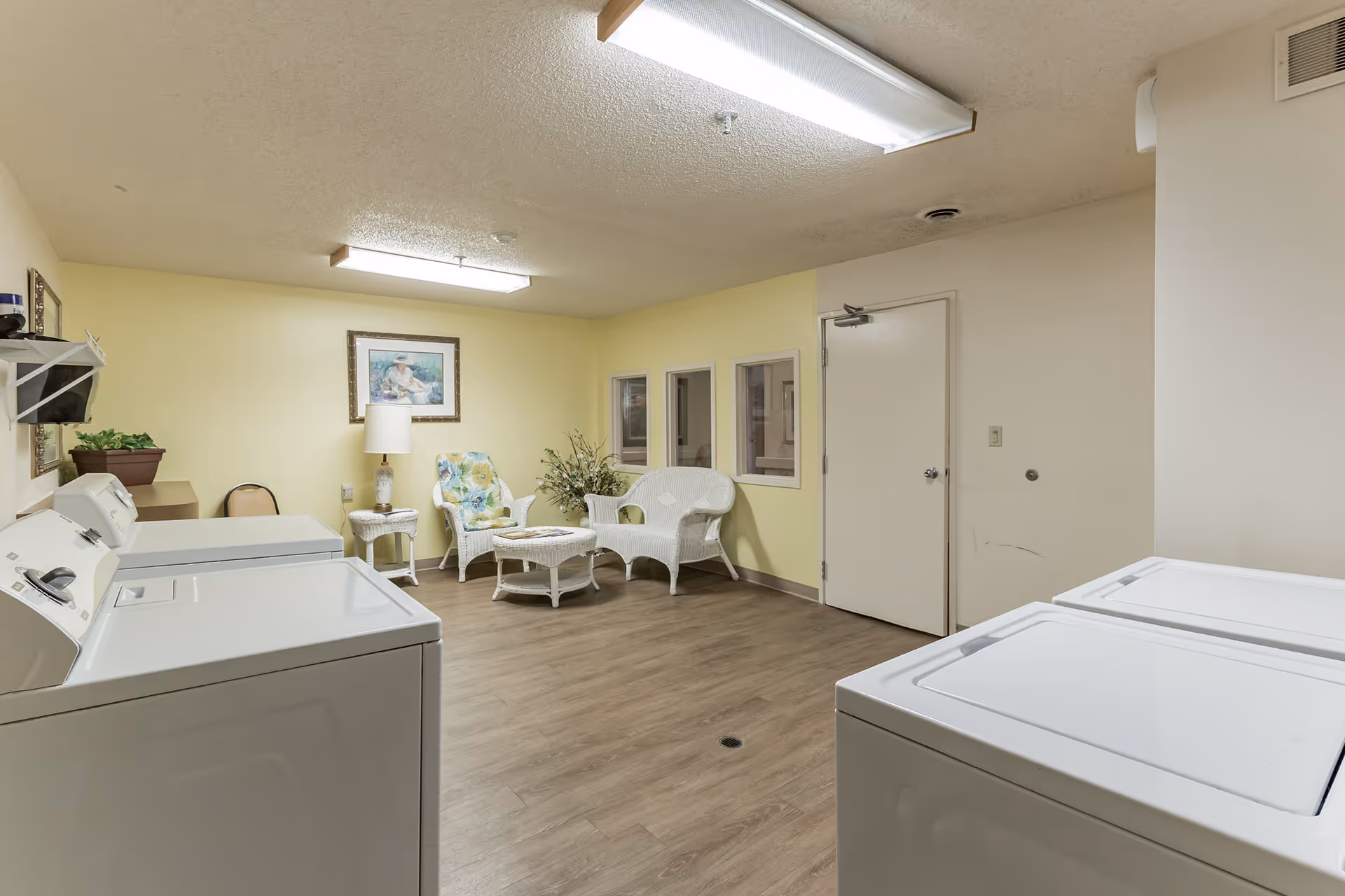 Laundry room with four white washing machines and dryers, a seating area with white wicker furniture including a chair, loveseat, and table, a lamp, a framed painting on a yellow wall, and wood flooring.