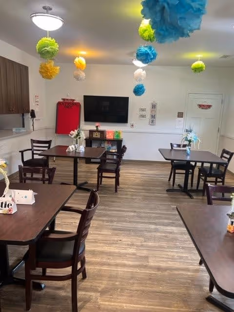 A dining room with several dark wood tables and chairs arranged on a wood floor. Colorful paper pom-pom decorations in blue, green, yellow, and white hang from the ceiling. There is a flat-screen TV mounted on the far wall above a small shelf with decorative items. The room has white walls and a door on the right side. Flower arrangements are placed on the tables.