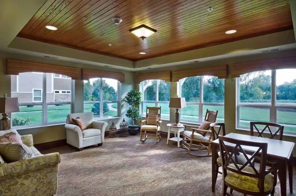 A bright and cozy living room area with large windows offering a view of green outdoor space. The room features a wooden ceiling with recessed lighting, several cushioned armchairs, rocking chairs, a small side table with a lamp, and a dining table with chairs. The space is carpeted and decorated with a potted plant and window valances.