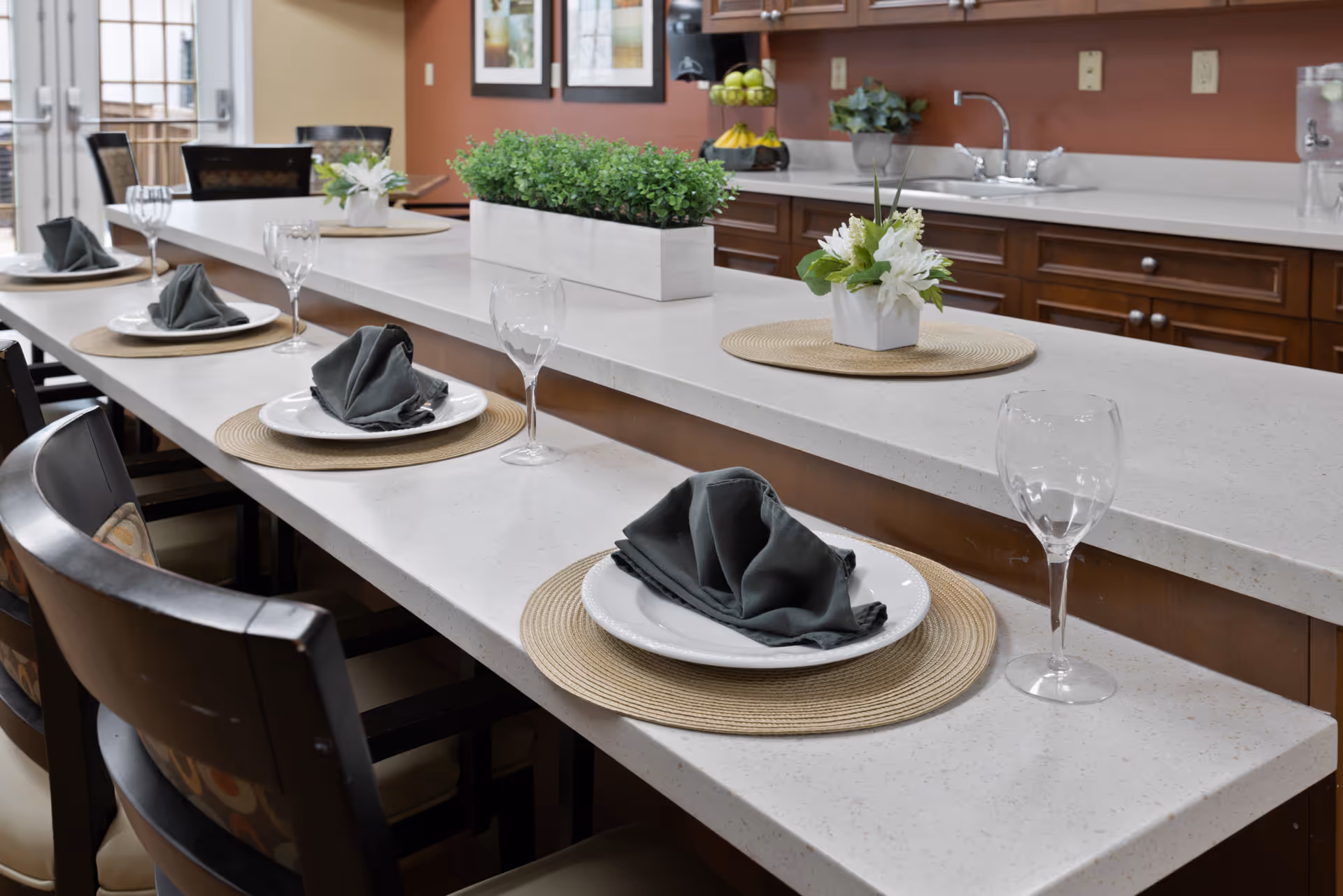 A modern kitchen counter set for dining with white plates, gray folded napkins, and empty wine glasses on round woven placemats. The counter has a white surface with a rectangular planter of green plants and a small floral arrangement. Dark wooden cabinets and a sink are visible in the background.