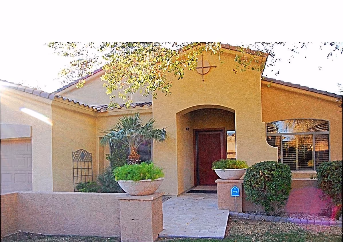 Front exterior view of a single-story assisted living home with beige stucco walls, a red front door, two large planters with green plants on either side of the entrance, a small palm tree, and neatly trimmed bushes. The building has a tiled roof and a paved walkway leading to the door.