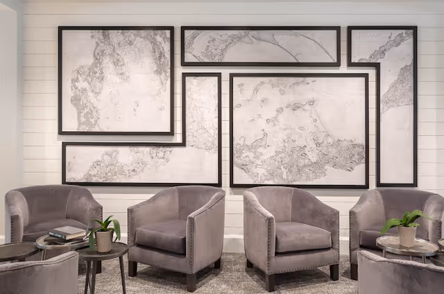 A cozy seating area with four gray upholstered armchairs arranged around two small round tables. Each table has a potted plant, and one table also has a stack of books. The background features a white shiplap wall decorated with multiple framed abstract art pieces in black and white.