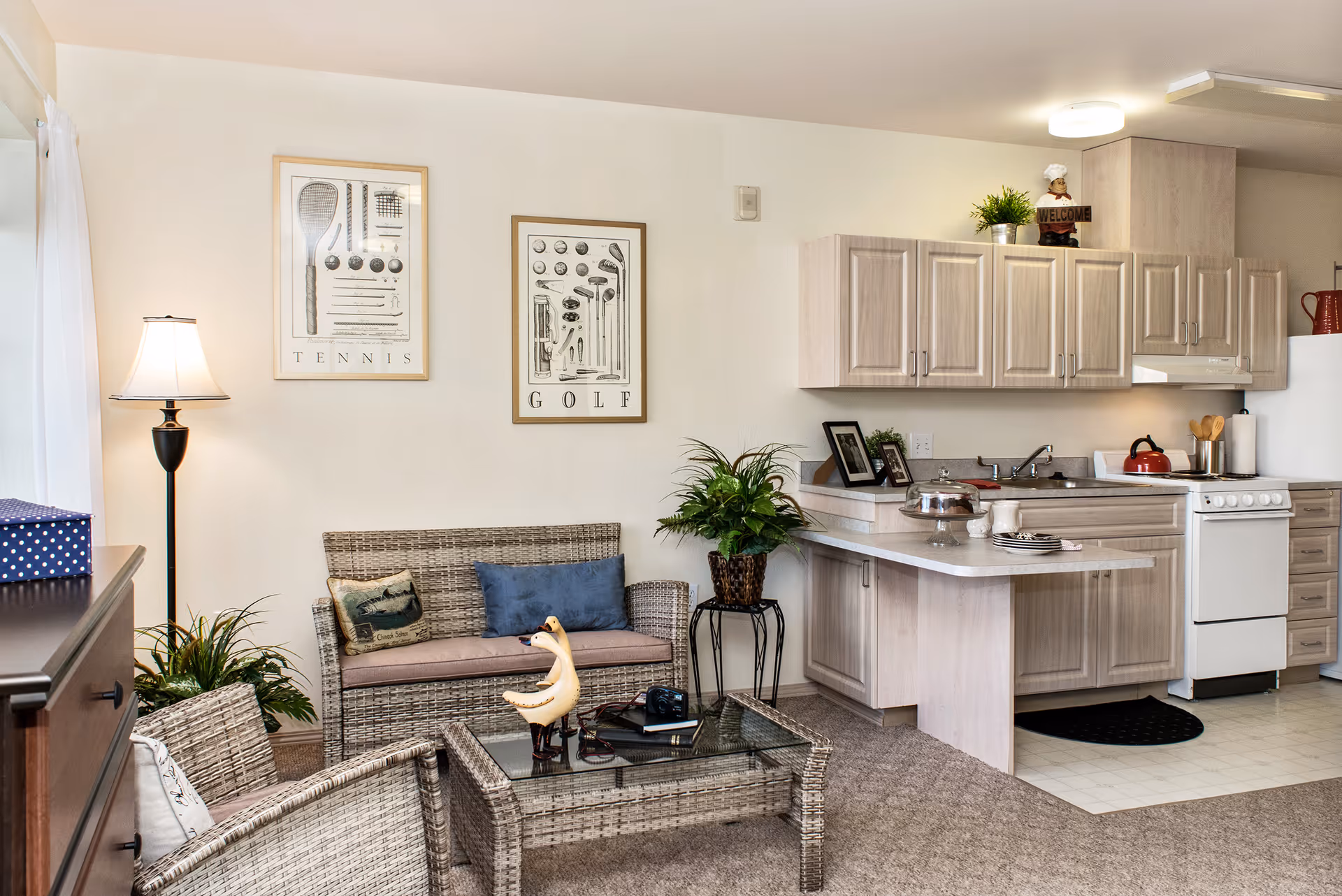 A cozy living area and kitchen in a continuing care community. The living area features wicker furniture including a loveseat and two chairs with cushions, a glass-top coffee table with decorative items, and a floor lamp. The kitchen has light wood cabinets, a white stove, a refrigerator, and a countertop with plates and a cake stand. Two framed sports-themed prints labeled 'Tennis' and 'Golf' hang on the wall above the loveseat. There are also some potted plants adding greenery to the space.