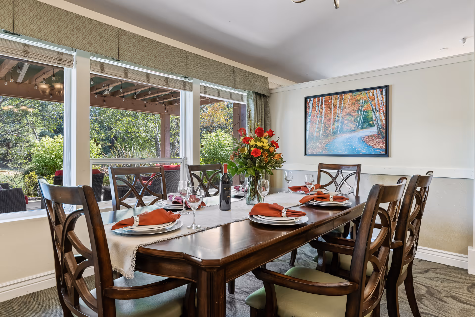 A dining room with a wooden table set for six people. The table has a white runner, plates with orange napkins in silver napkin rings, wine glasses, a bottle of red wine, and a vase with a colorful flower arrangement. Large windows show an outdoor patio with greenery and string lights. A framed picture of a forest path with autumn foliage hangs on the wall.